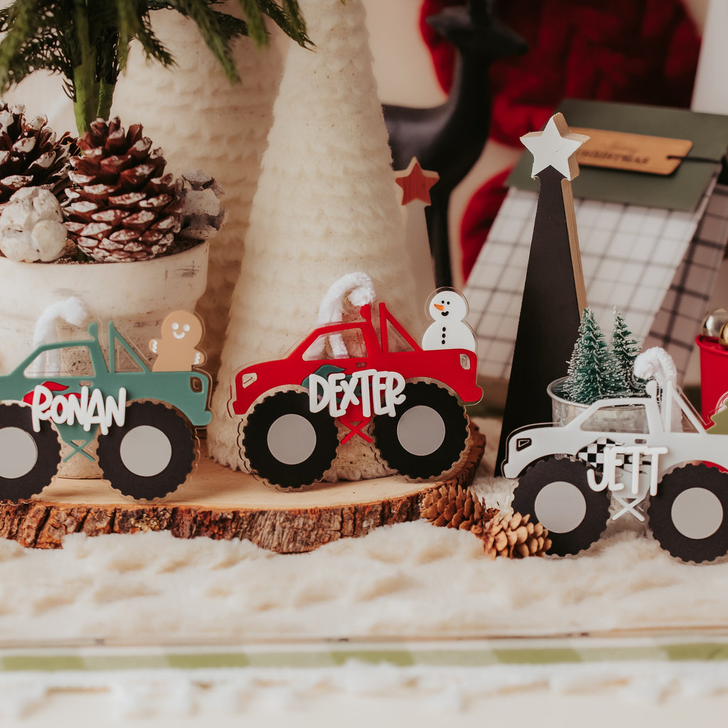 Three personalized acrylic monster truck ornaments displayed on a rustic wood slice, decorated with Christmas-themed props like pinecones, a small tree, and holiday decorations in the background.