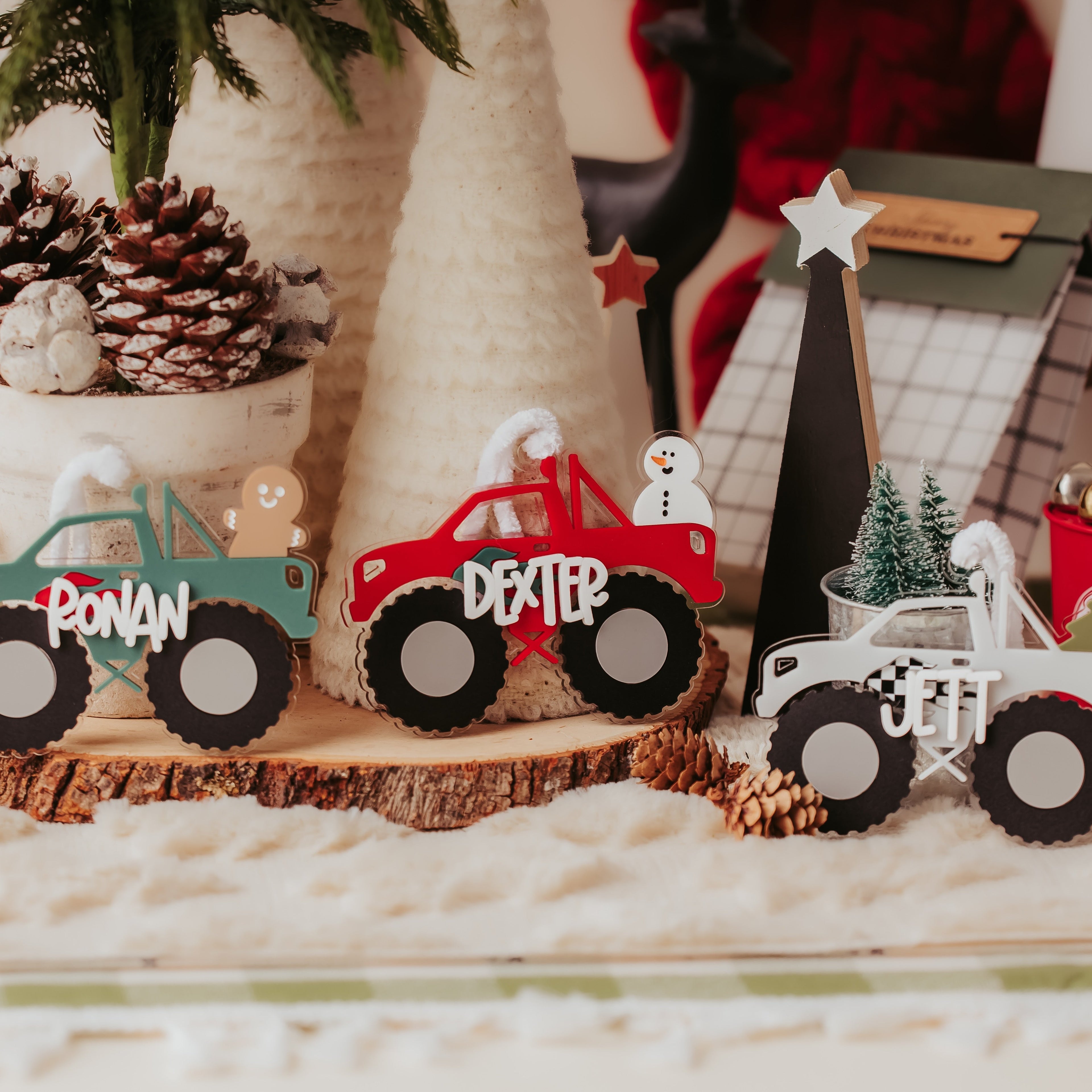 Three personalized acrylic monster truck ornaments displayed on a rustic wood slice, decorated with Christmas-themed props like pinecones, a small tree, and holiday decorations in the background.
