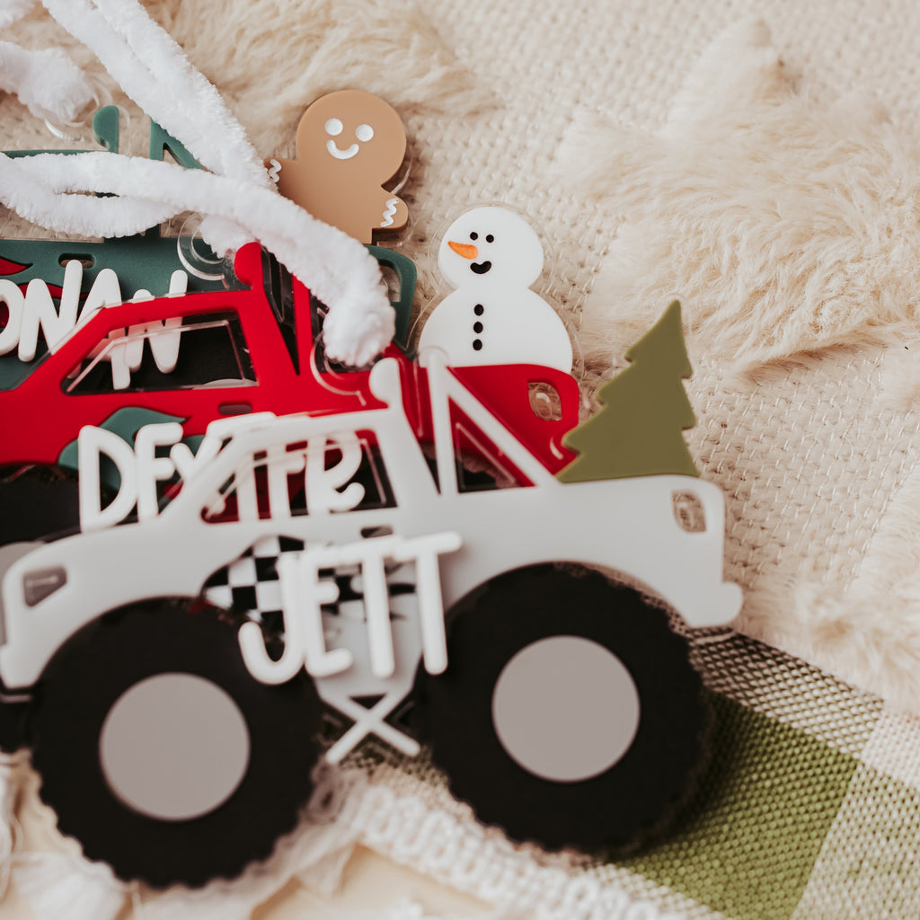 A close-up of three Christmas monster truck ornaments, a red truck carrying a white snowman and a green Christmas tree, with a gingerbread man figure and white lettering, placed on a textured cream fabric.