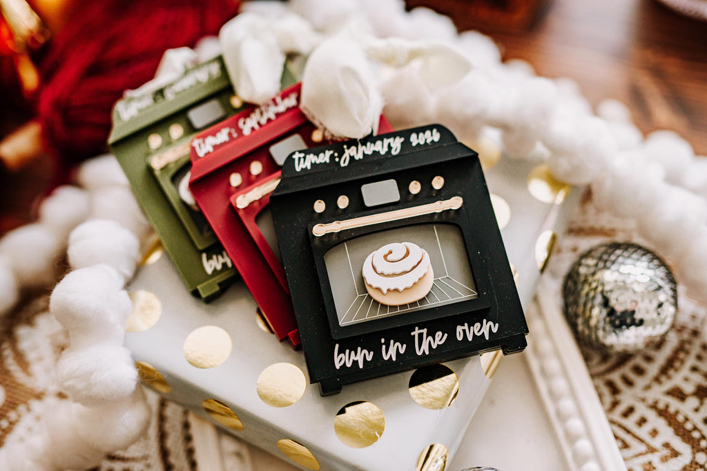A set of christmas ornaments shaped like mini ovens, displayed on a white gift box with gold polka dots. The front tag is black with white script reading 'timer: January 2025' and 'bun in the oven' below, showing a cinnamon roll graphic.