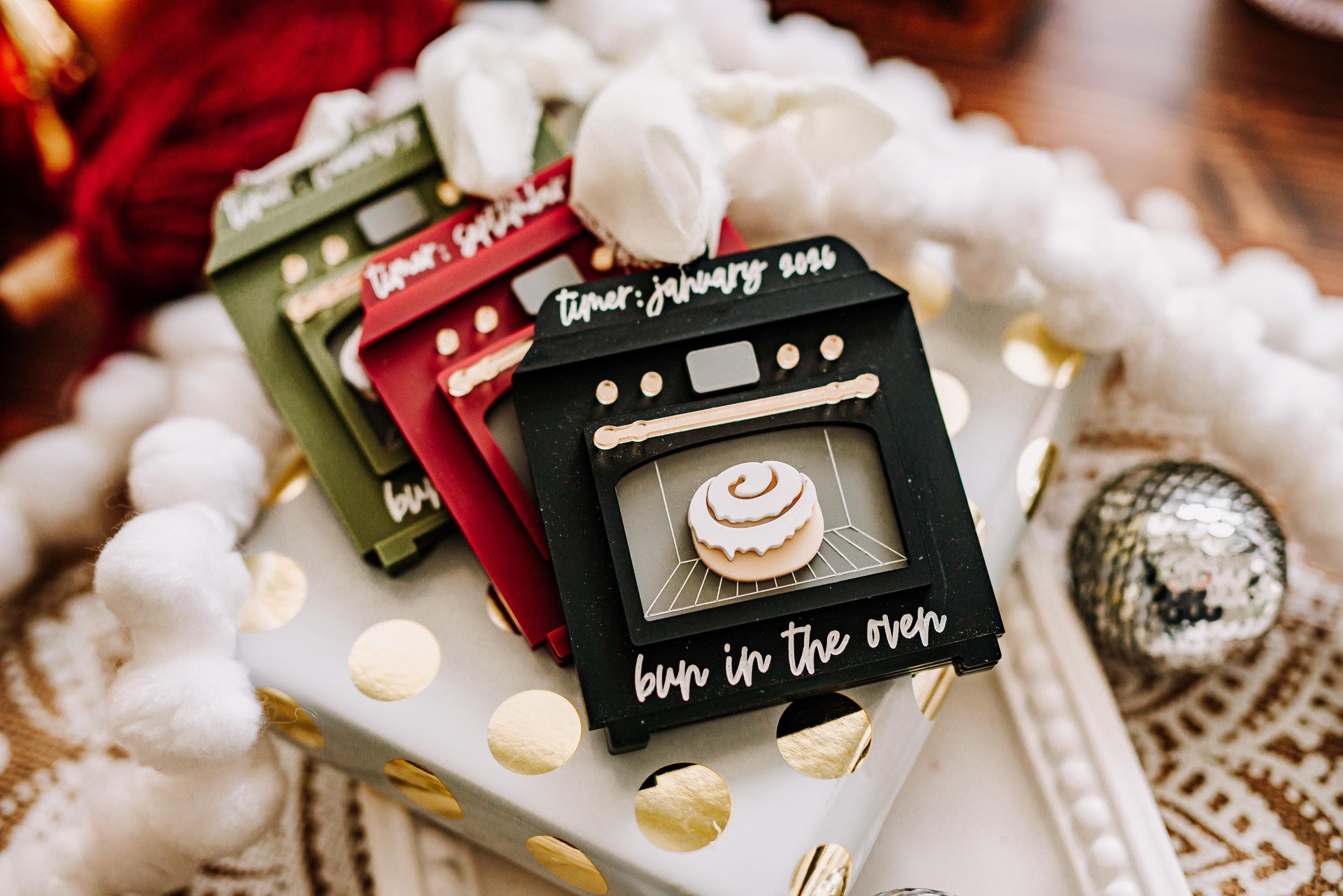A set of christmas ornaments shaped like mini ovens, displayed on a white gift box with gold polka dots. The front tag is black with white script reading 'timer: January 2025' and 'bun in the oven' below, showing a cinnamon roll graphic.