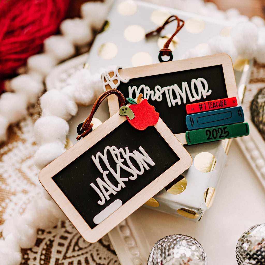 Two chalkboard Christmas ornaments on a cream and gold colored gift box. One chalkboard ornament has an apple with a bite missing and a worm coming out of it. The other chalkboard ornament has a stack of books with the year and 'ABC' at the top.