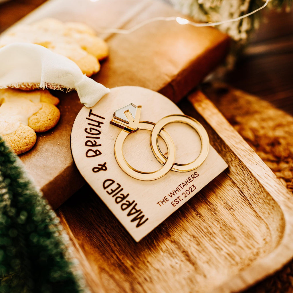 A personalized wooden arch shaped wedding ornament with engraved text reading 'Married & Bright' and 'The Whitakers Est. 2023,' featuring a gold-tone 3d interlocking rings, resting on a wooden tray with holiday greenery and warm lighting.