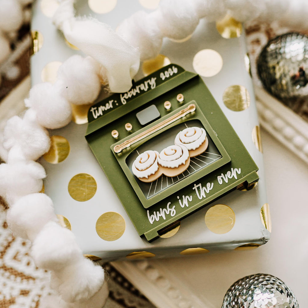 A styled gift box wrapped in white paper with gold polka dots, featuring a pregnancy announcement  ornament that reads 'buns in the oven' with an illustration of three iced cupcakes. The box is decorated with white pom-pom garlands and surrounded by holiday ornaments on a patterned surface. 