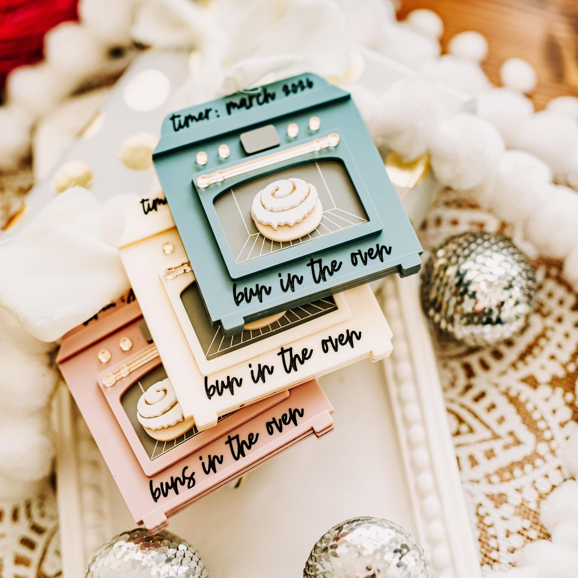 Three small decorative oven-shaped ornaments on a white tray, with the top piece in teal reading 'timer: march 2015' and the lower pieces in cream and blush pink. The scene is styled with white pom-pom garland, silver disco balls, and a warm wooden surface in the background.