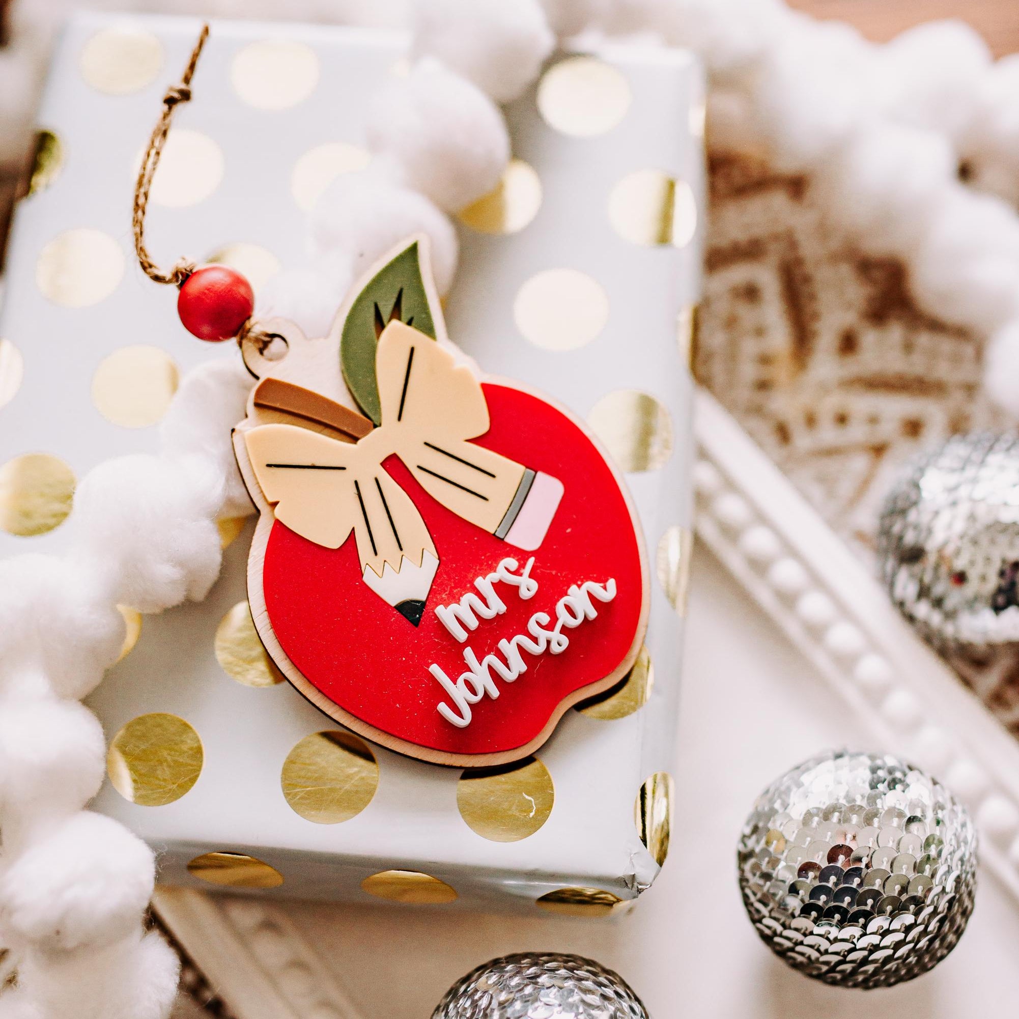 A red apple-shaped teacher Christmas ornament with a gold bow and the words 'Mrs. Johnson' in white script, resting on a wrapped gift box with gold polka dots