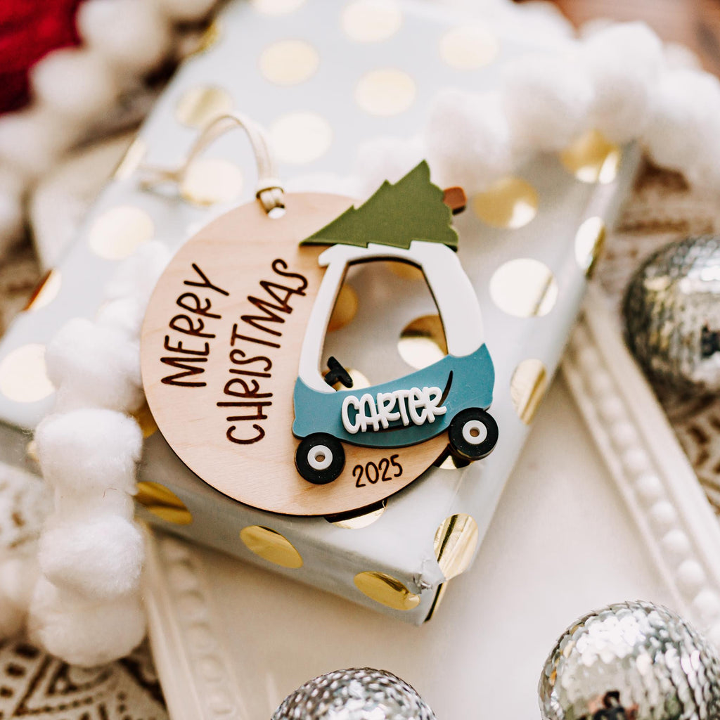 A personalized Christmas ornament shaped like a cozy coupe  car with a tree on top, hanging on a gift box wrapped in white and gold polka dot paper. The ornament reads 'Merry Christmas' and 'Carter 2025'.