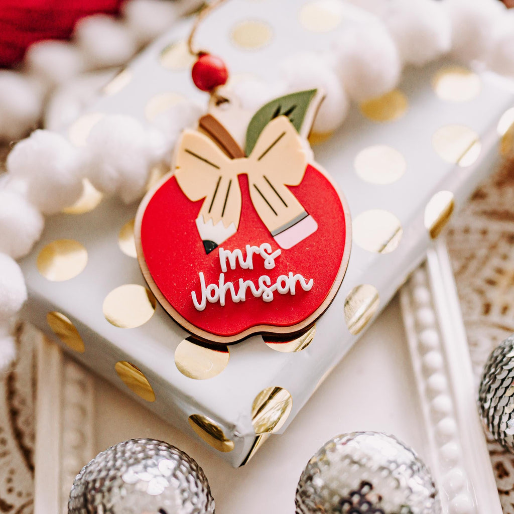 A close-up of a red apple-shaped ornament with a yellow pencil shaped bow and the words 'Mrs. Johnson' in white script, placed on a white gift box with gold polka dots and surrounded by white pom-pom garland and silver ornaments.