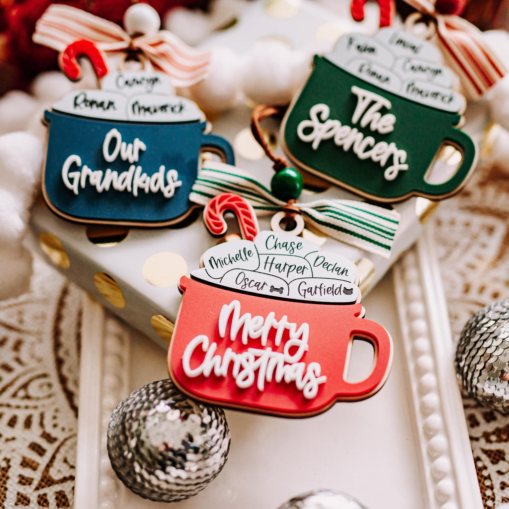 Three Christmas hot cocoa mug ornaments with humorous names like 'Our Grandkids' and 'The Spencers' and 'Merry Christmas' written on them, styled on a gift box with white pom-pom beads and silver ornaments.