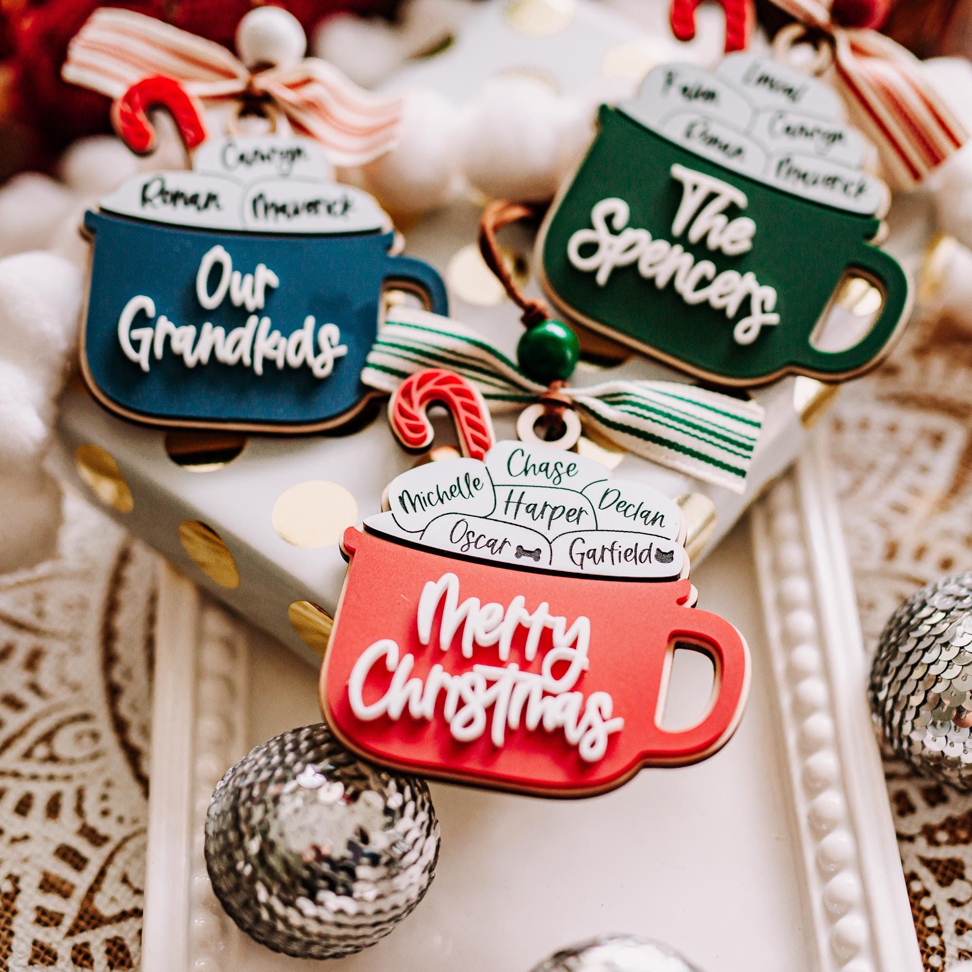 Three Christmas hot cocoa mug ornaments with humorous names like 'Our Grandkids' and 'The Spencers' and 'Merry Christmas' written on them, styled on a gift box with white pom-pom beads and silver ornaments.