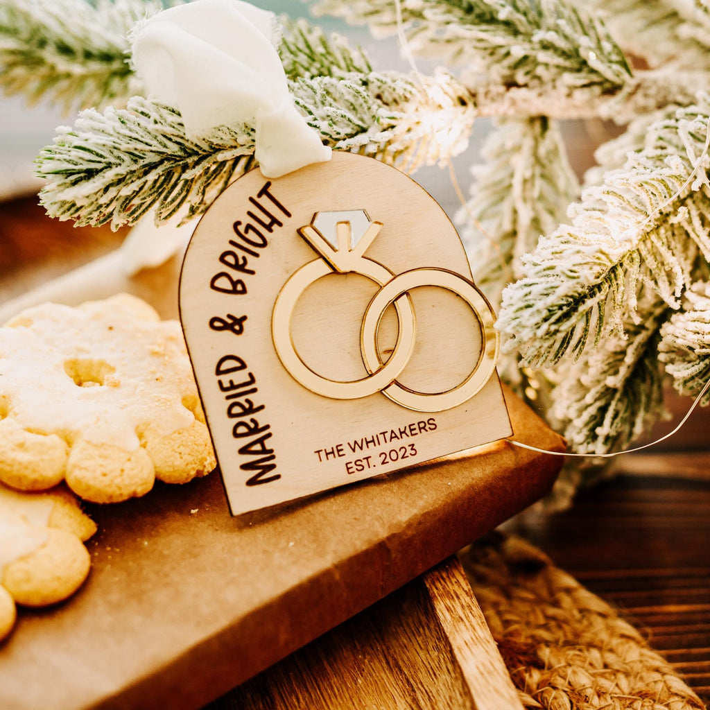 A personalized wooden arch Christmas ornament with 3D gold interlocking rings and engraved with  'MARRIED & BRIGHT' and 'THE WHITAKER EST. 2025', hanging near frosted pine branches and cookies on a wooden board.