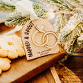 A personalized wooden arch Christmas ornament with 3D gold interlocking rings and engraved with  'MARRIED & BRIGHT' and 'THE WHITAKER EST. 2025', hanging near frosted pine branches and cookies on a wooden board.